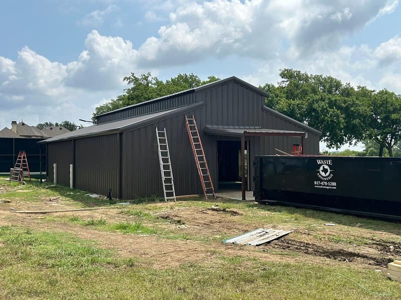 Dark brown metal shop with two ladders set at the eave and a dumpster alongside