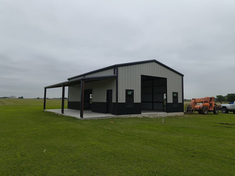Gray and black metal shop with a covered side porch, seen from the corner across the lawn