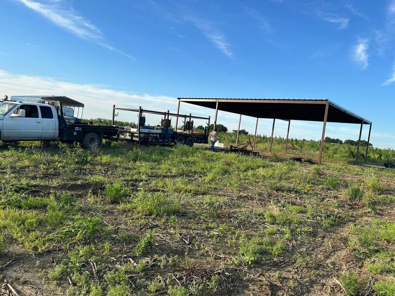 Open-sided metal hay cover on red steel posts, pickup and trailer parked beside it