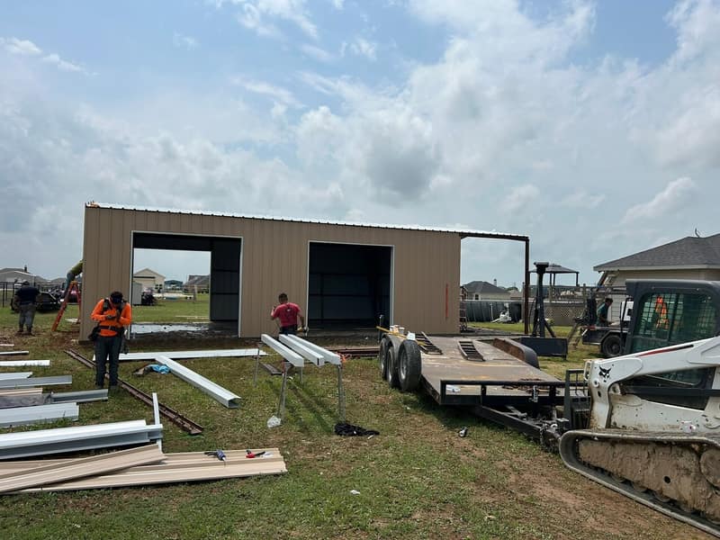 Crew setting steel beside a tan metal shop with two bay openings, trailer and skid steer on site