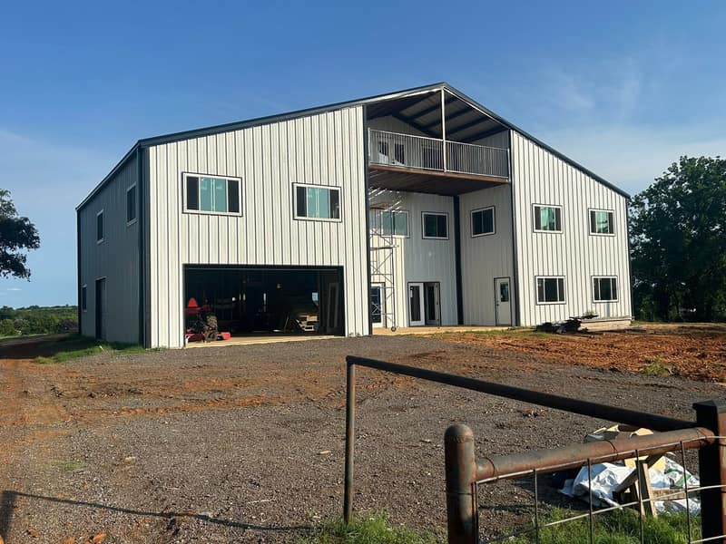 Two-story white metal barndominium with a balcony, an open garage bay and a pipe fence in front