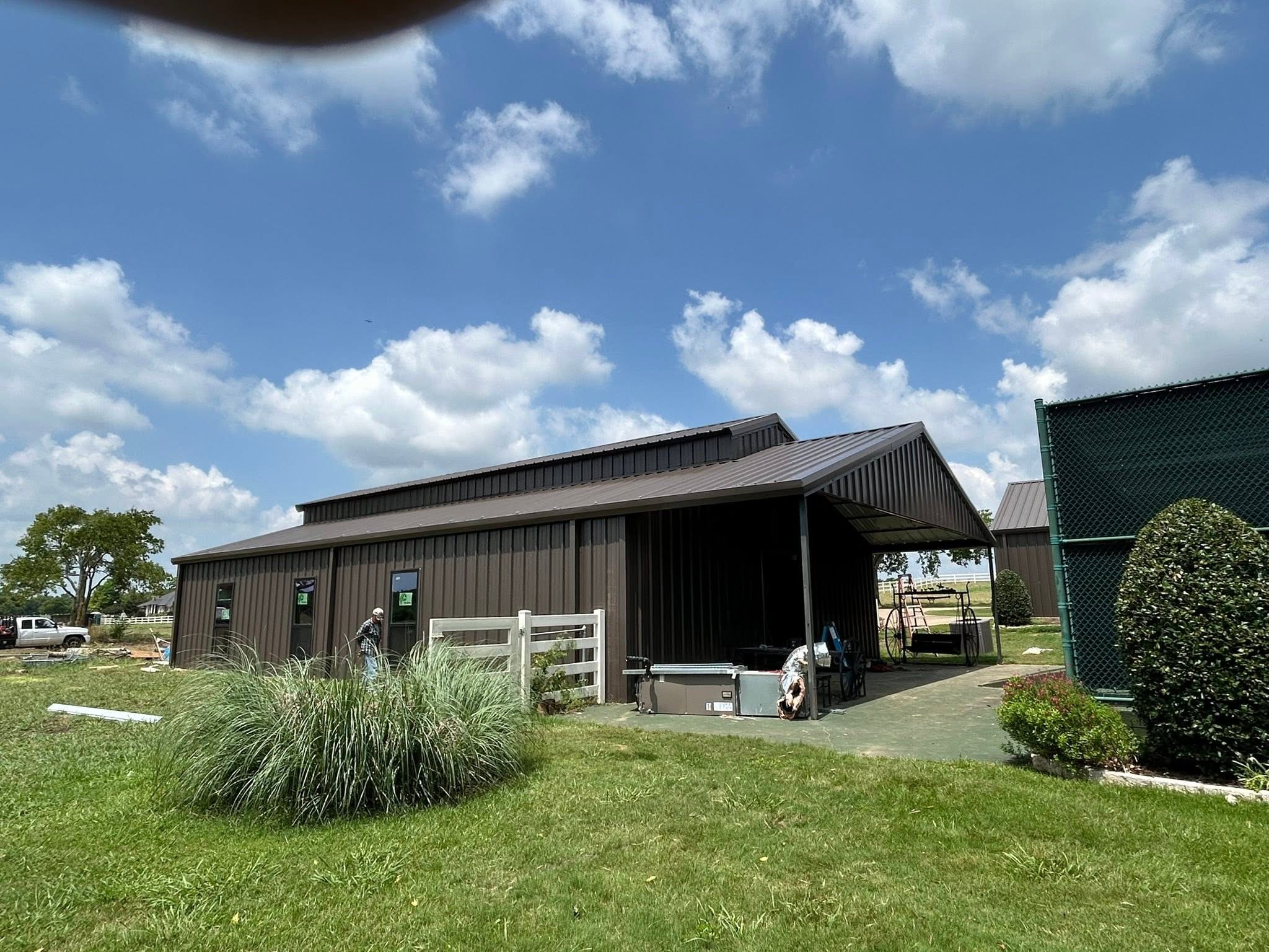 Dark brown metal barn with a covered side patio and white pipe fencing